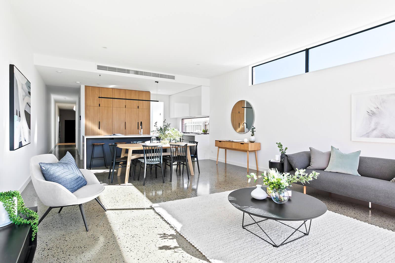 Light filled living room with property styling. Gray toned lounge, armchair and coffee table. Timber kitchen cupboards complemented with timber dining table and console. Green and white floral arrangements.