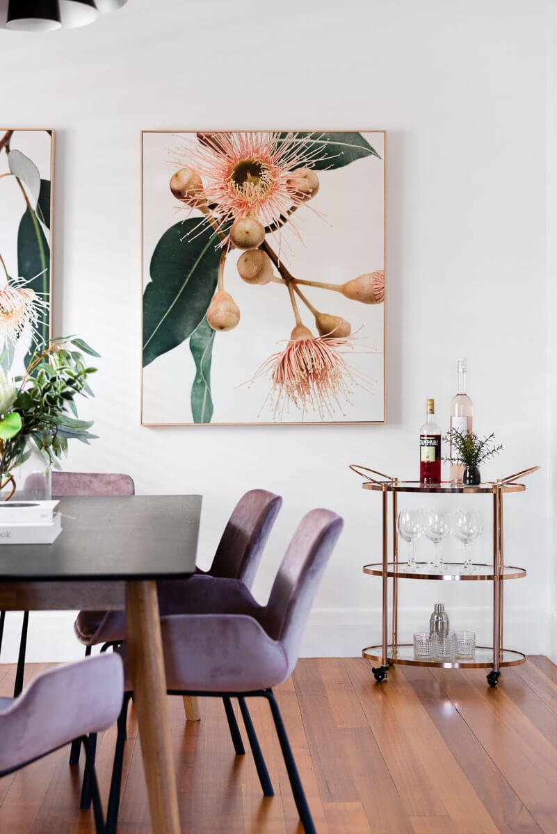 Formal living room with modern mauve velvet dining chairs around a black rectangular dining table. Also shows a large print with gum tree flowers above a copper and glass drinks trolley.