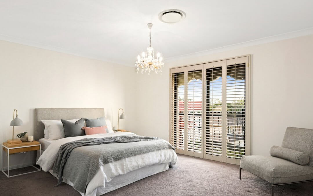 Chermside West Master Bedroom styled with queen bed, soft pink and grey accessories. The bed is framed by matching timber and white framed bedside tables. A grey occasional chair sits in a corner opposite the bed.