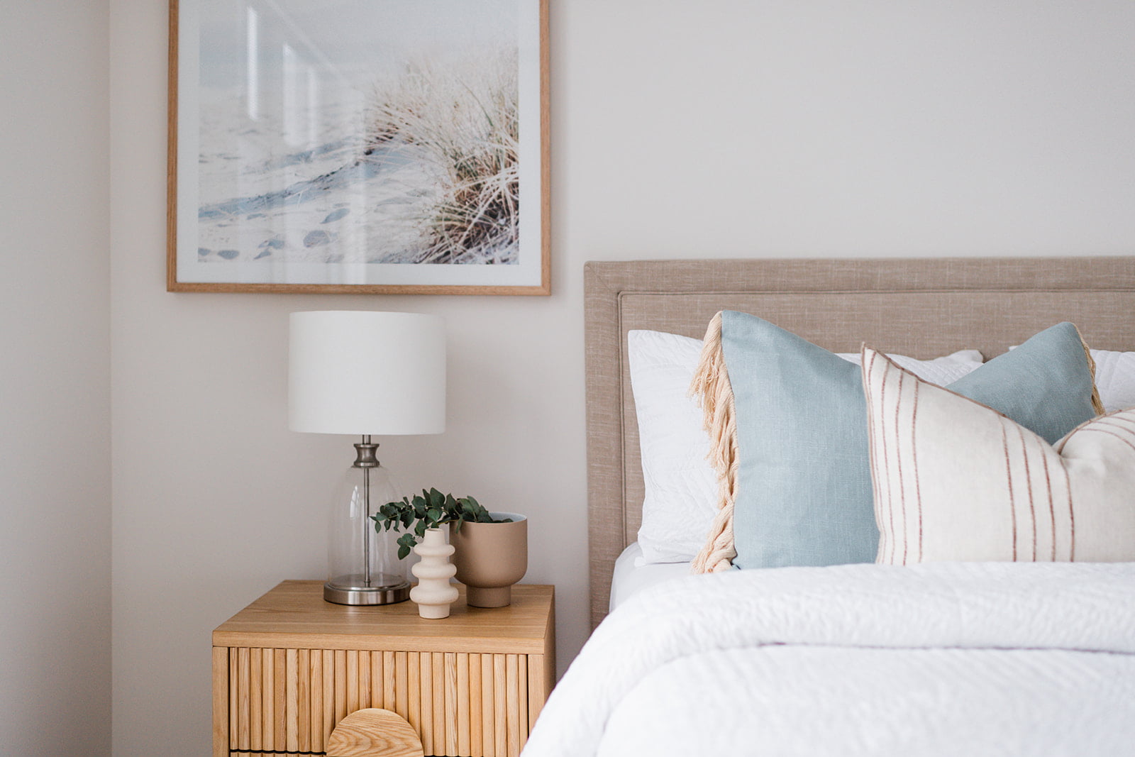 Bed with pink cushions, grey throw, white linen. Timber bedside tables with woven white table lamps. Grey bedhead with centred artwork above with hues of pink, blue and navy.